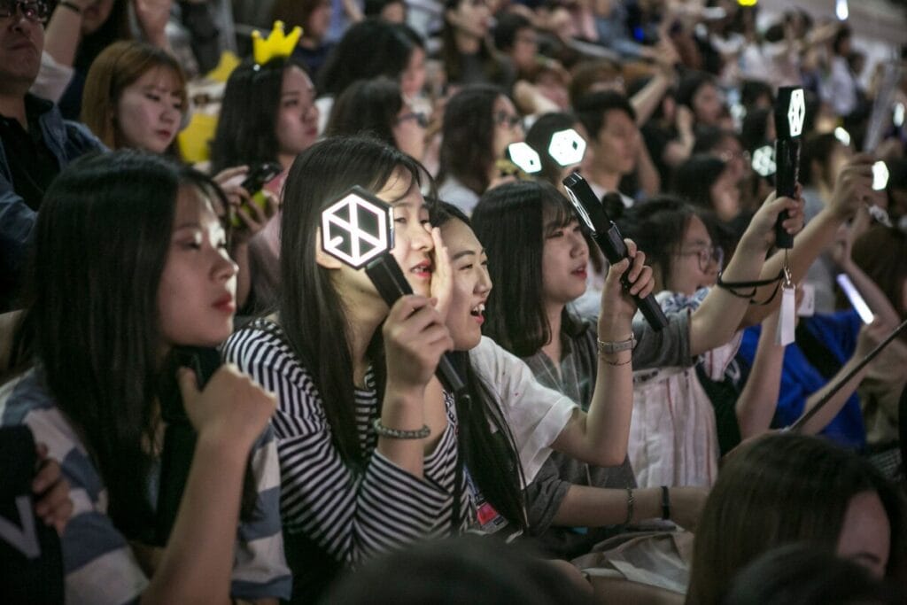 A diverse crowd of young fans at a K-pop concert. Several young women in the foreground are holding up and looking at glowing K-pop light sticks and a smartphone, with other fans visible in the background. The fans are a multi-ethnic group in a dark concert environment.