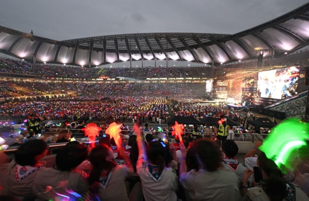 A wide shot of a massive, brightly lit stadium concert at night. In the foreground, fans are seen from behind holding glowing red light sticks, with a huge, crowded stadium and bright stage screens in the background.